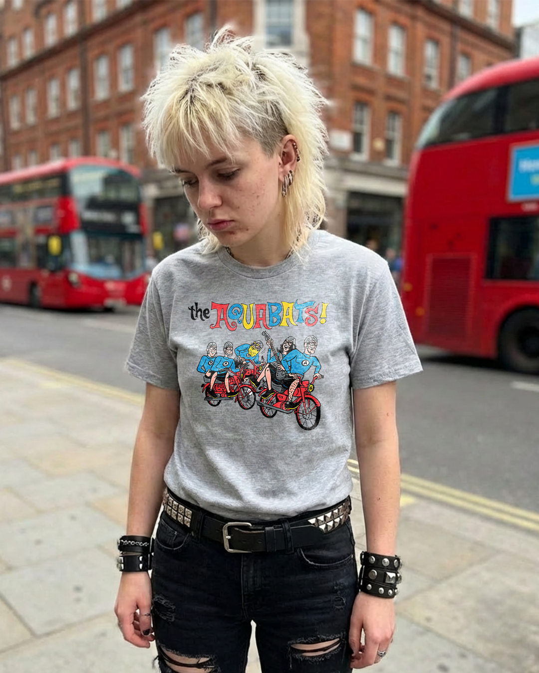 Person wearing a gray t-shirt with 'The Aquabats!' graphic on a street with red double-decker buses.